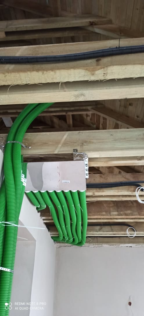 Zielone, karbowane rury wentylacyjne przymocowane do drewnianej konstrukcji sufitu poddasza, widoczna skrzynka rozdzielcza i czarne przewody.