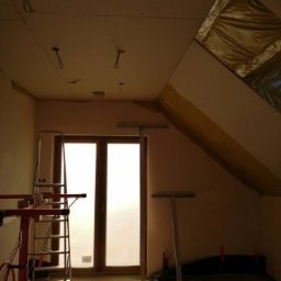 Grzegorz Idziak Anowi Usługi remontowo-budowlane - Poddasze w trakcie prac wykończeniowych: zamontowane płyty gipsowo-kartonowe na skosach, widoczne profile, drabina i narzędzia malarskie, okno zasłonięte folią, przygotowane punkty oświetleniowe.