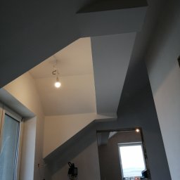Grzegorz Idziak Anowi Usługi remontowo-budowlane - Wnętrze w trakcie malowania: szare ściany i sufit w geometrycznym układzie, widoczna żarówka na kablu, okno w tle i narzędzia malarskie.