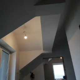 Wnętrze w trakcie malowania: szare ściany i sufit w geometrycznym układzie, widoczna żarówka na kablu, okno w tle i narzędzia malarskie.