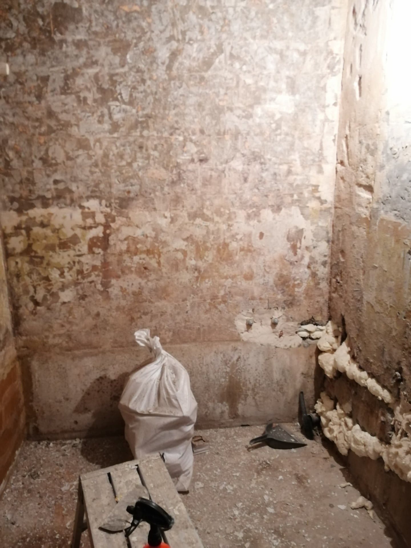 Wnętrze pomieszczenia w trakcie przygotowania do prac wykończeniowych; widoczne ślady starej farby na ceglanej ścianie, częściowo usunięte tynki, pianka montażowa w rogu pomieszczenia, worek...