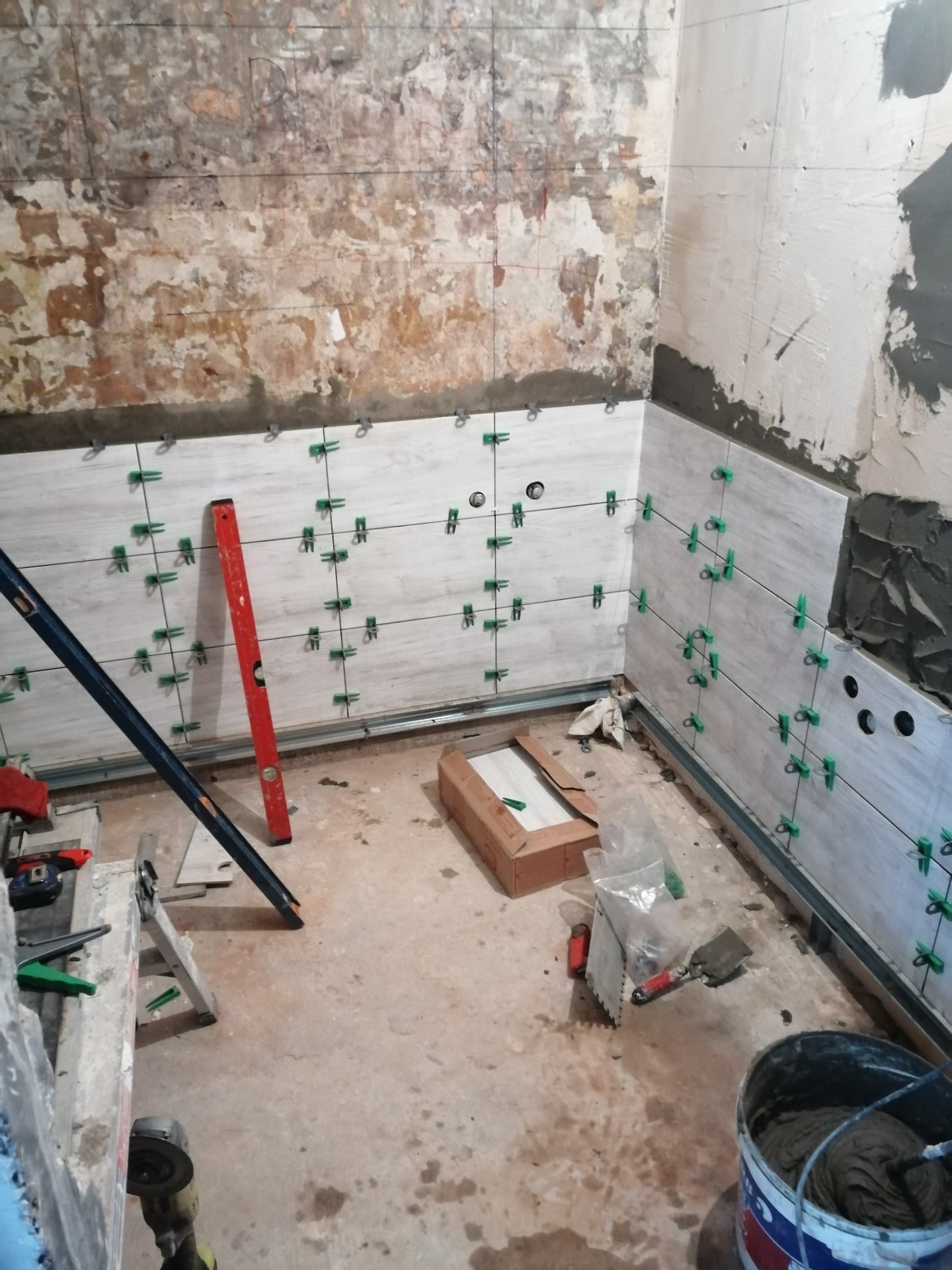 Ściana łazienki w trakcie układania jasnych płytek imitujących drewno, widoczne zielone klipsy poziomujące, poziomica i narzędzia glazurnicze, ściana częściowo z surowym tynkiem.