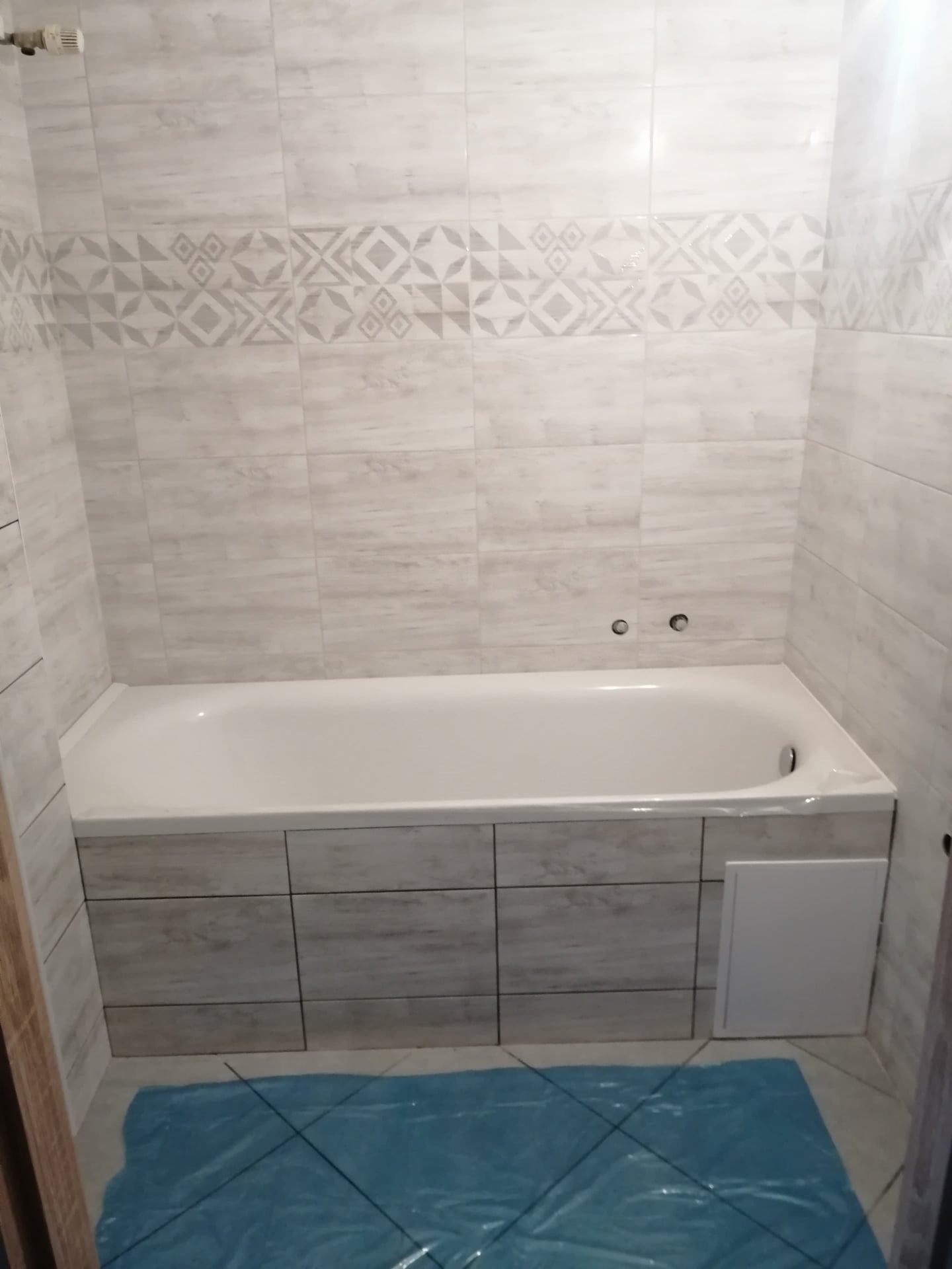 Wykończona łazienka z wanną obudowaną płytkami imitującymi drewno, ściany wyłożone jasnymi płytkami z dekoracyjnym pasem z geometrycznym wzorem, podłoga częściowo zakryta niebieską folią ochronną.