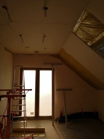 Poddasze w trakcie prac wykończeniowych: zamontowane płyty gipsowo-kartonowe na skosach, widoczne profile, drabina i narzędzia malarskie, okno zasłonięte folią, przygotowane punkty oświetleniowe.