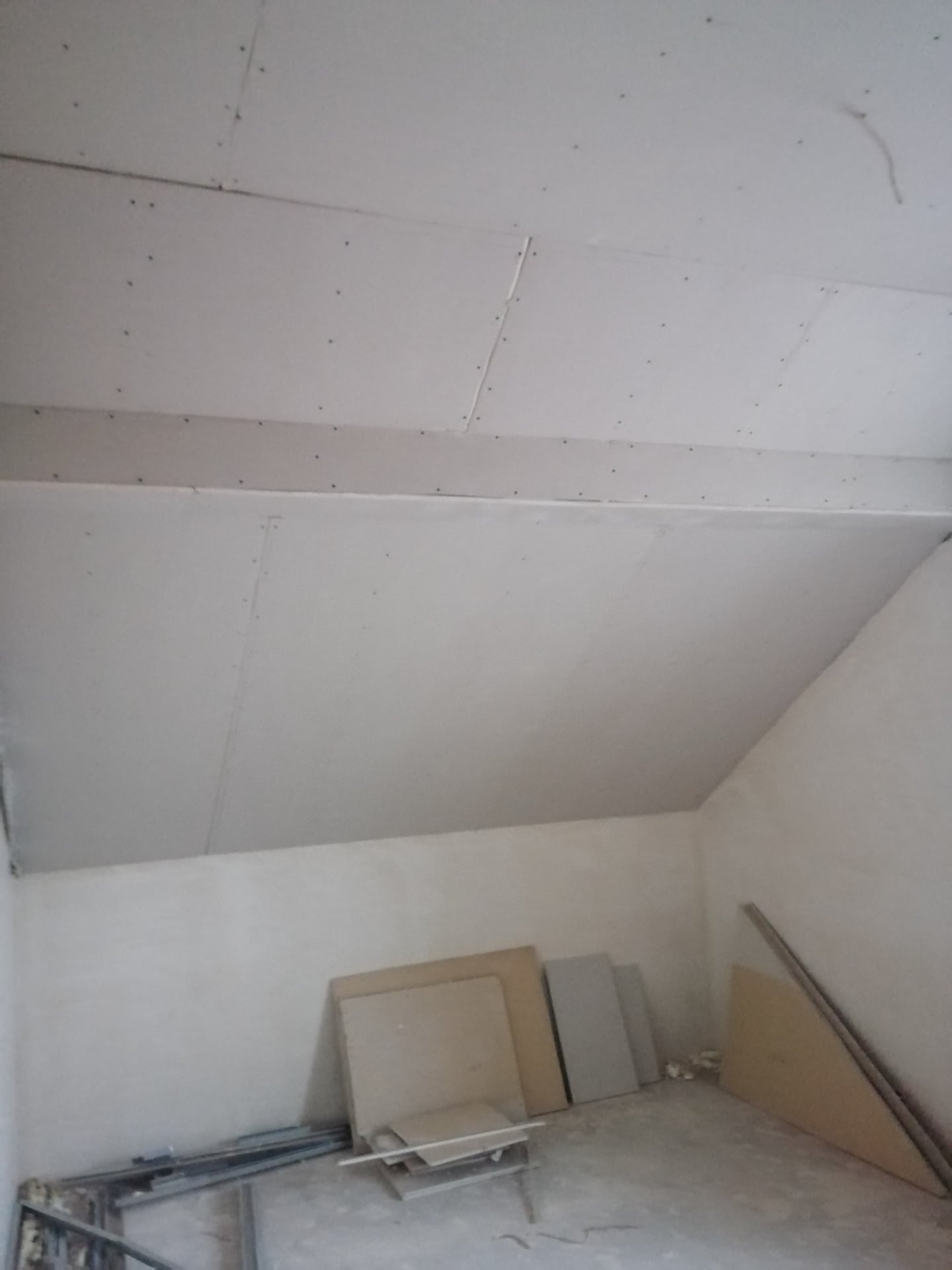 Sufit poddasza pokryty płytami gipsowo-kartonowymi, widoczne wkręty, w tle ściana i płyty gipsowe o różnych rozmiarach opierające się o ścianę.