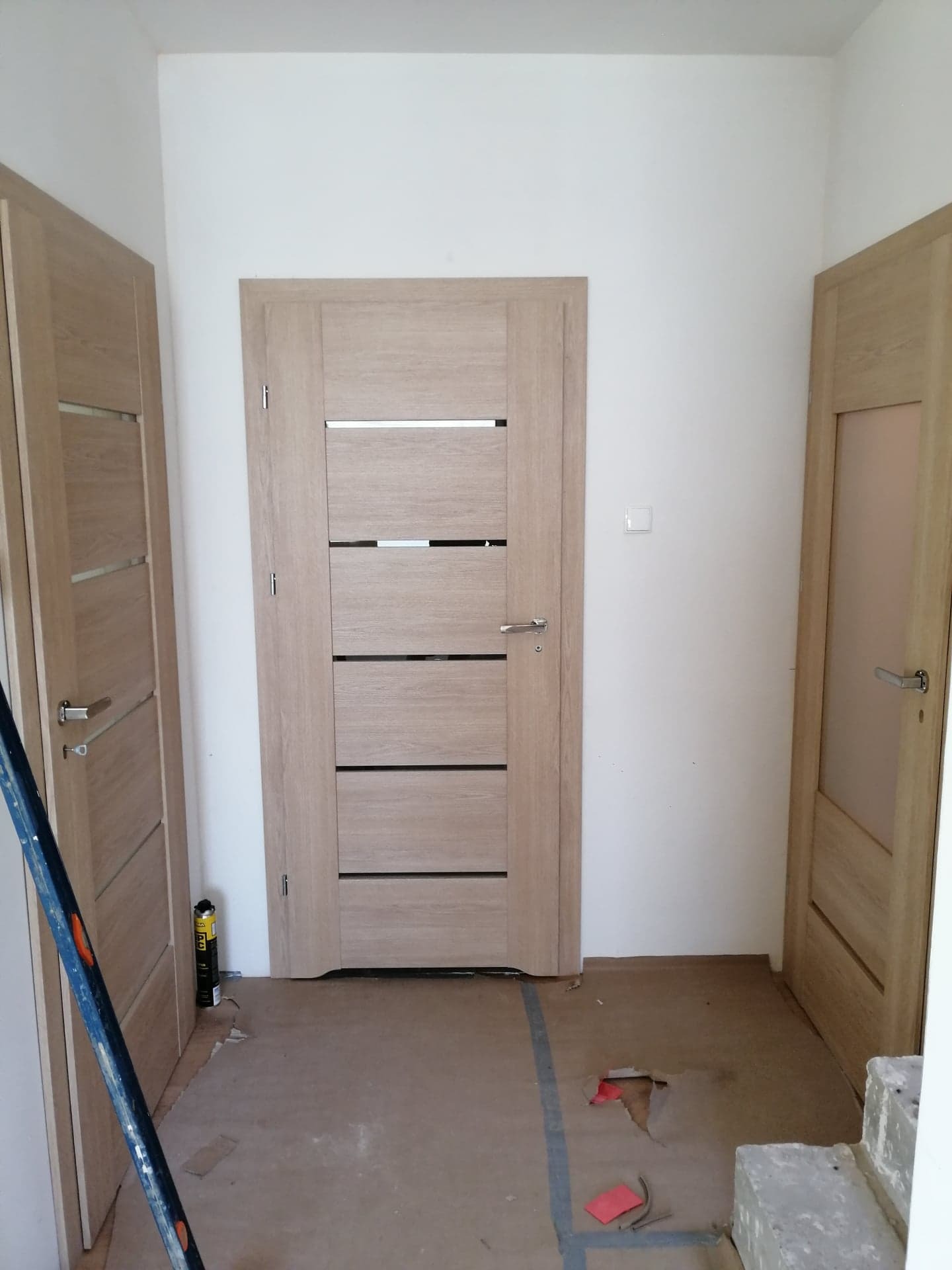 Montaż trzech wewnętrznych drzwi z drewnianą okleiną o różnym designie, widoczne ślady prac na podłodze i puszka pianki montażowej.
