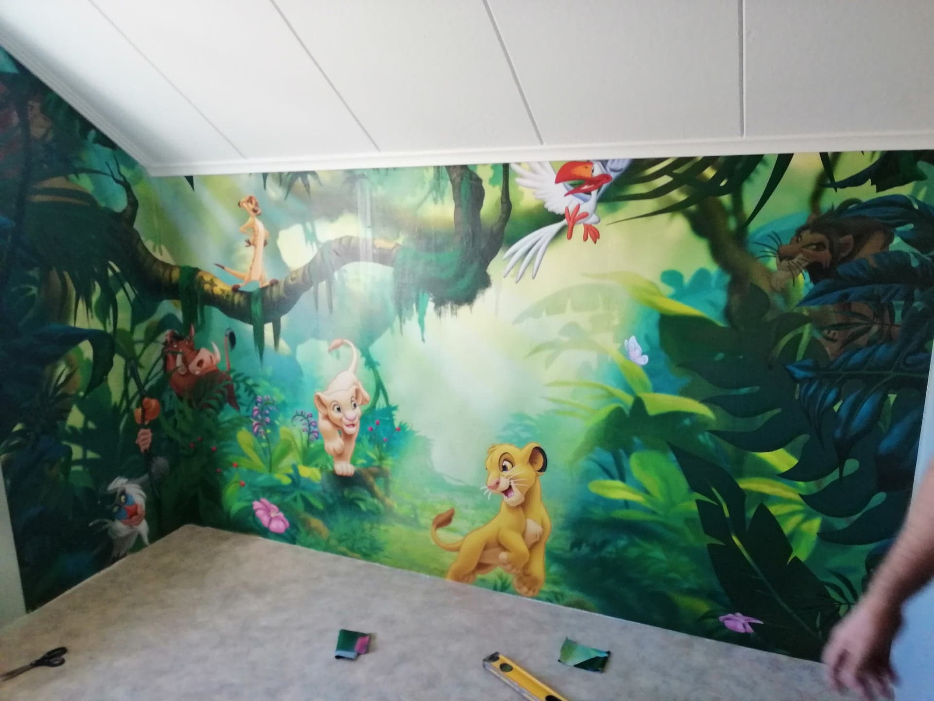 Ściana pokoju dziecięcego pokryta fototapetą z motywem Króla Lwa, widoczne narzędzia i fragment podłogi, praca w toku.