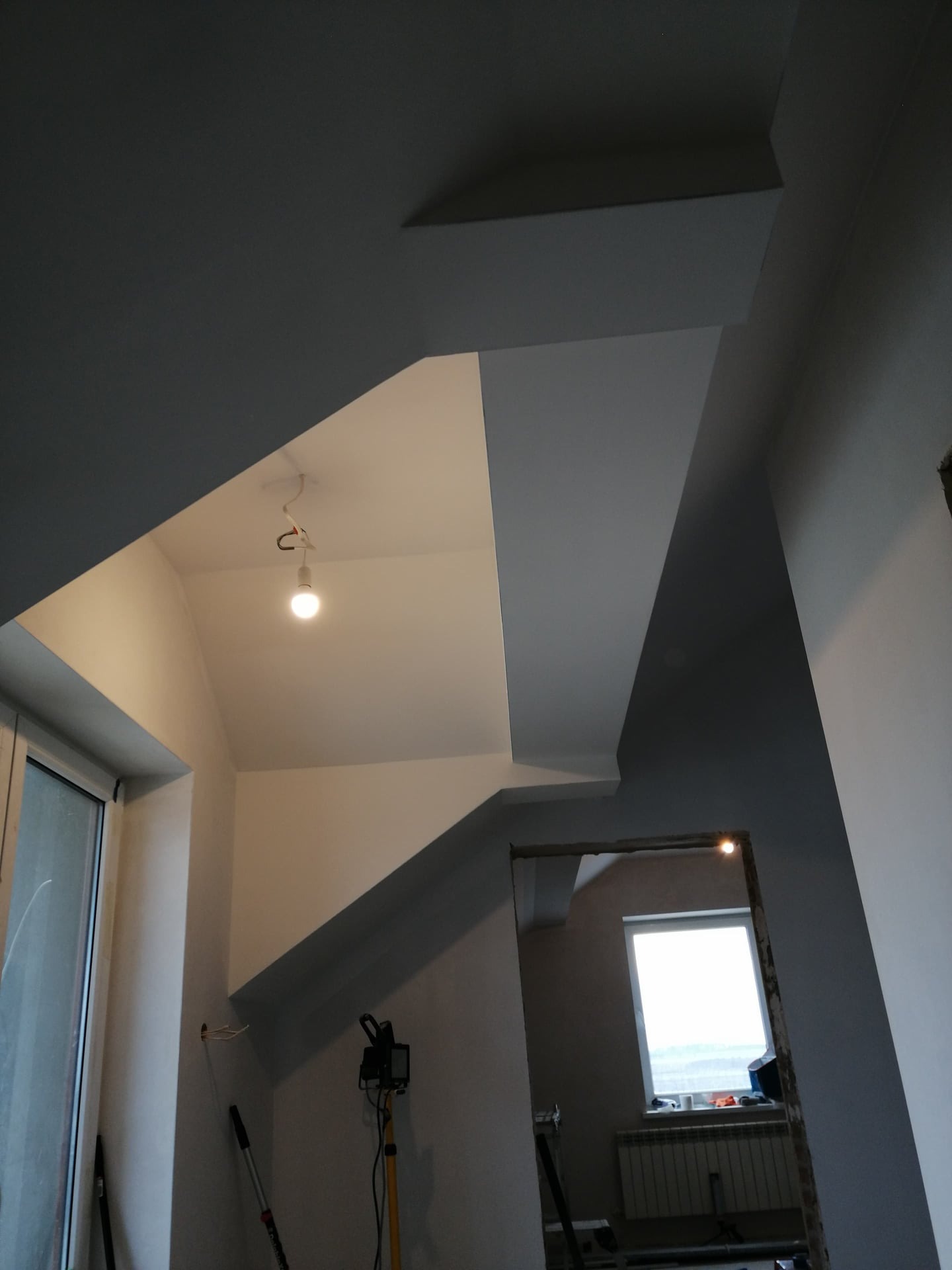Wnętrze w trakcie malowania: szare ściany i sufit w geometrycznym układzie, widoczna żarówka na kablu, okno w tle i narzędzia malarskie.