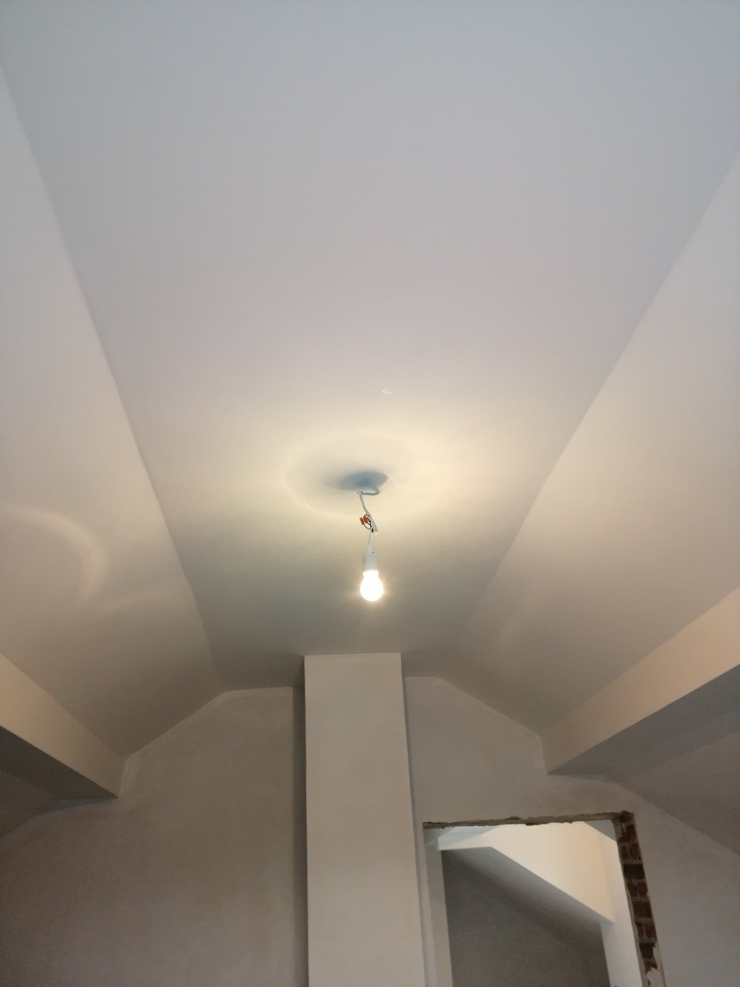 Świeżo wygładzony sufit z widocznymi łączeniami płyt gipsowo-kartonowych, przygotowany do malowania, oświetlony wiszącą żarówką bez klosza, w tle fragment ściany i otwór drzwiowy w trakcie remontu.