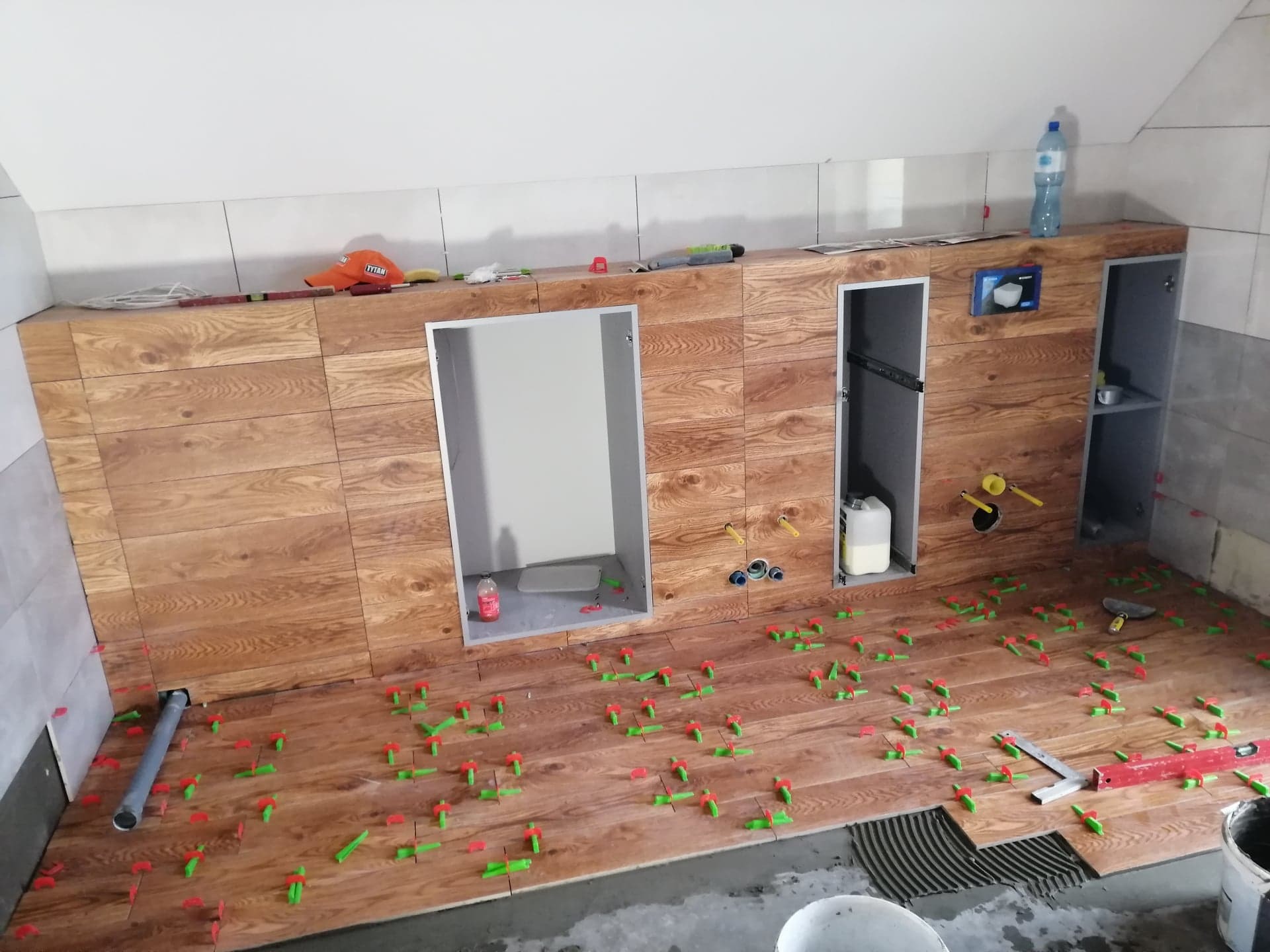 W trakcie układania płytek imitujących drewno w łazience, widoczne zielone i czerwone kliny poziomujące, przygotowane otwory na instalacje sanitarne, częściowo ułożona podłoga z widocznym klejem...