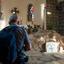 SacroPhoto Wioleta Żurawska - Mężczyzna z plecakiem i maseczką stoi w kościele Mensa Christi w Izraelu, patrząc na ołtarz z krzyżem i witraże.