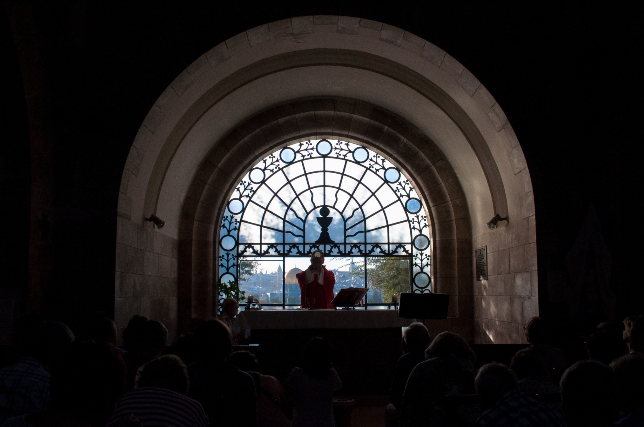 Wnętrze kościoła z wiernymi podczas mszy, ksiądz w czerwonej szacie przed oknem z dekoracyjnym witrażem, w tle widoczny krajobraz miejski.