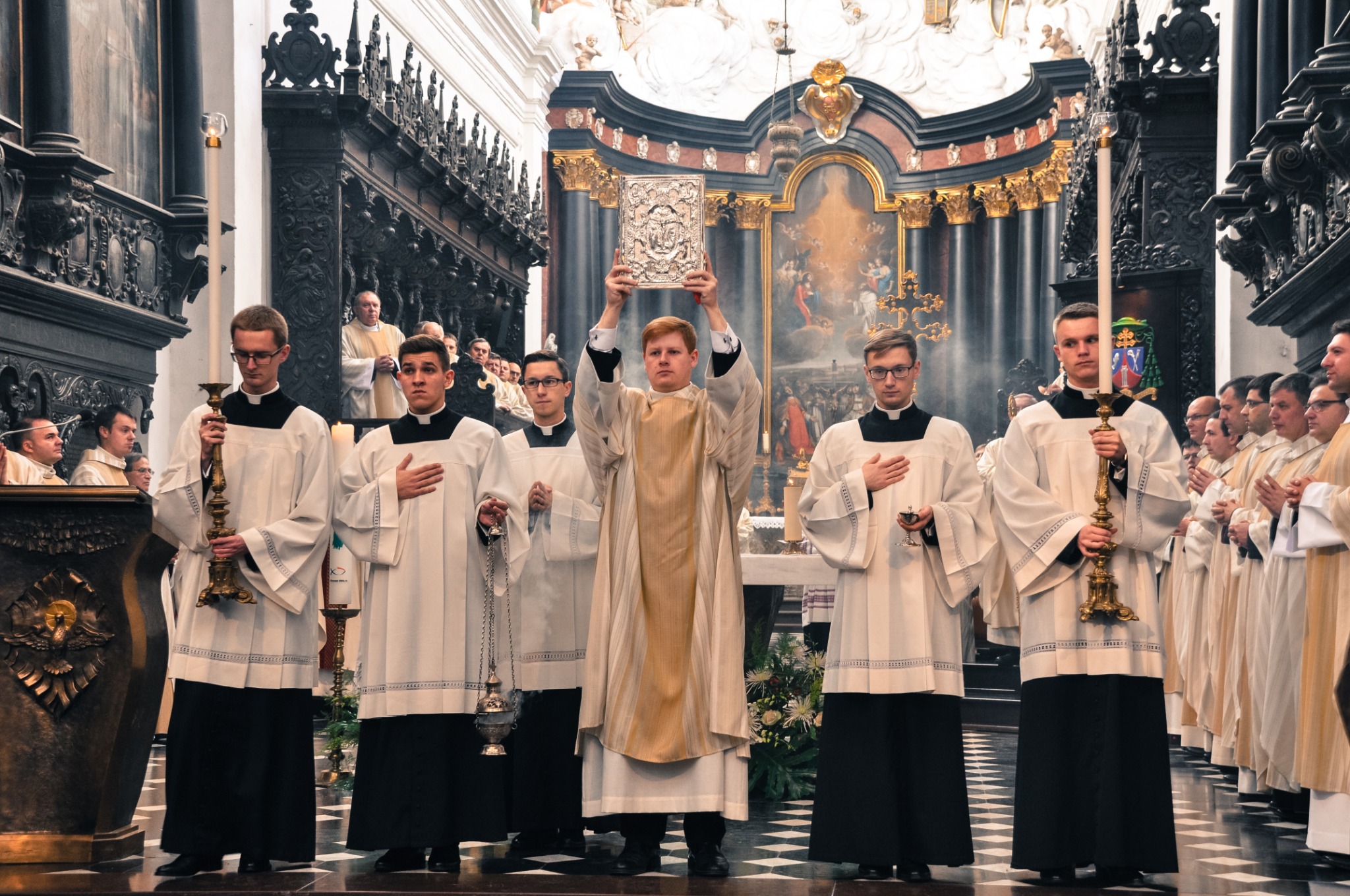 Wnętrze kościoła podczas uroczystości religijnej, grupa mężczyzn w białych szatach liturgicznych, jeden z nich trzyma nad głową bogato zdobioną księgę, w tle ołtarz i zgromadzeni wierni.