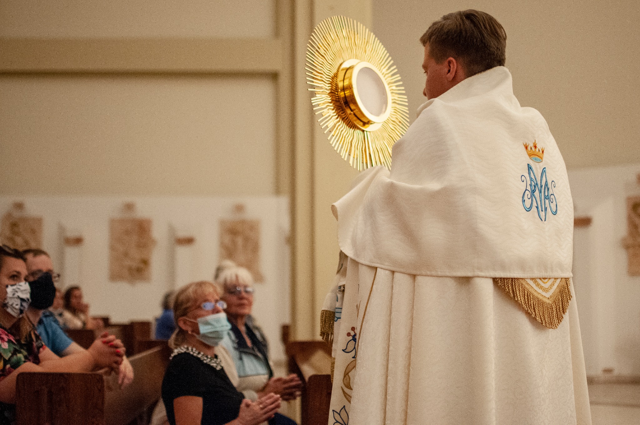 Ksiądz w ornacie z monstrancją podczas nabożeństwa, wierni w maseczkach siedzą w ławkach.
