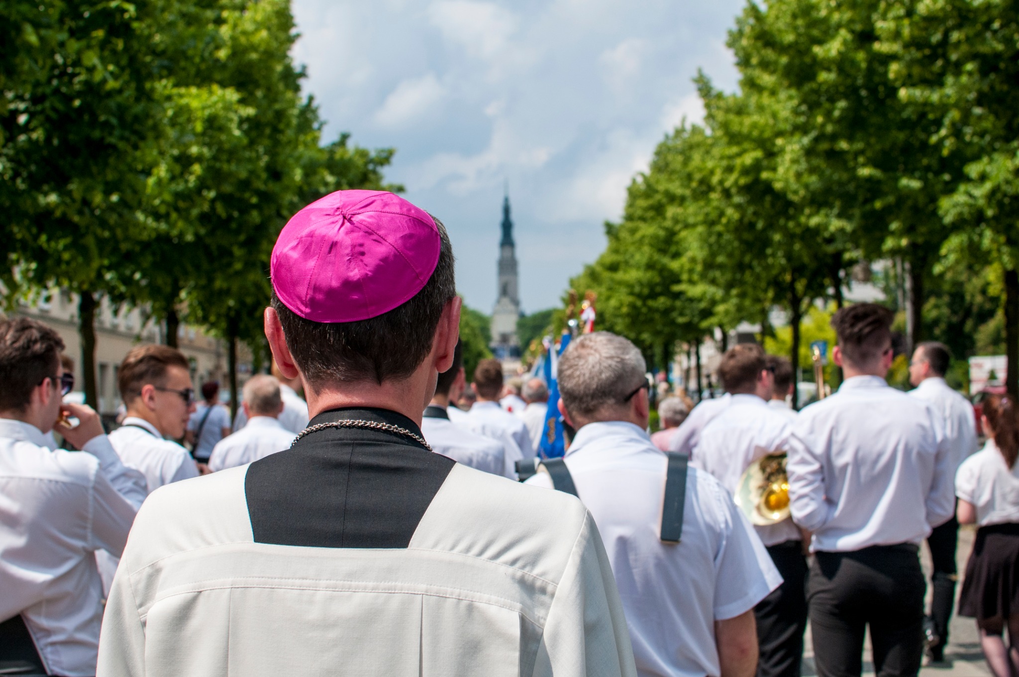 Procesja religijna z widokiem na wieżę kościoła, z perspektywy osoby w różowej piusce.
