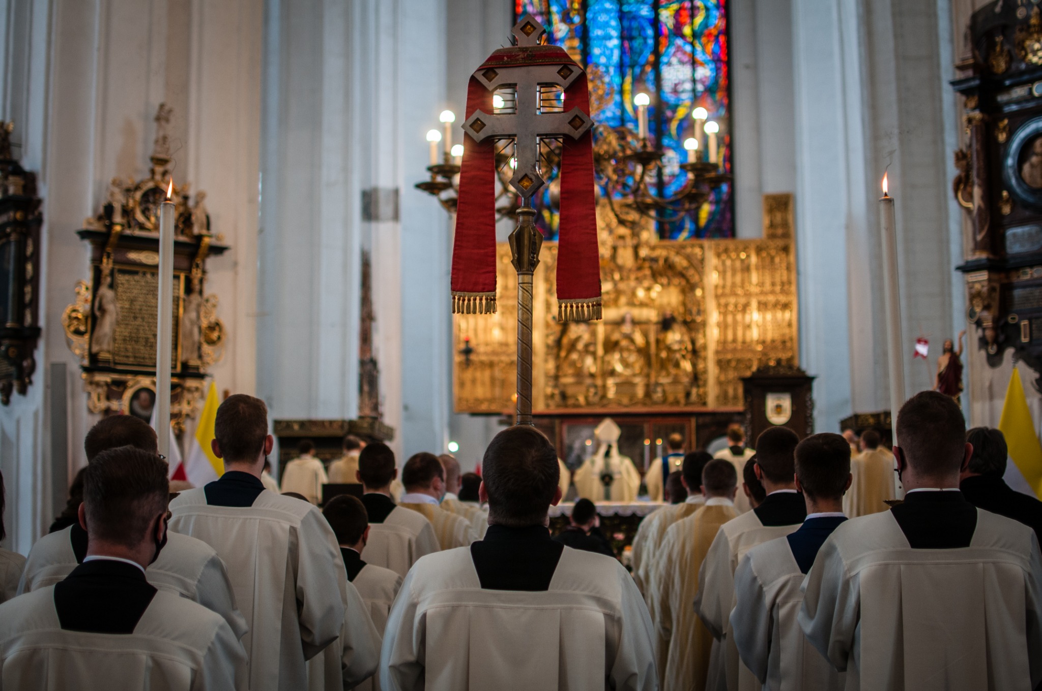 Wnętrze kościoła podczas uroczystości religijnej; widoczny procesyjny krzyż na pierwszym planie i zgromadzeni wierni w białych szatach
