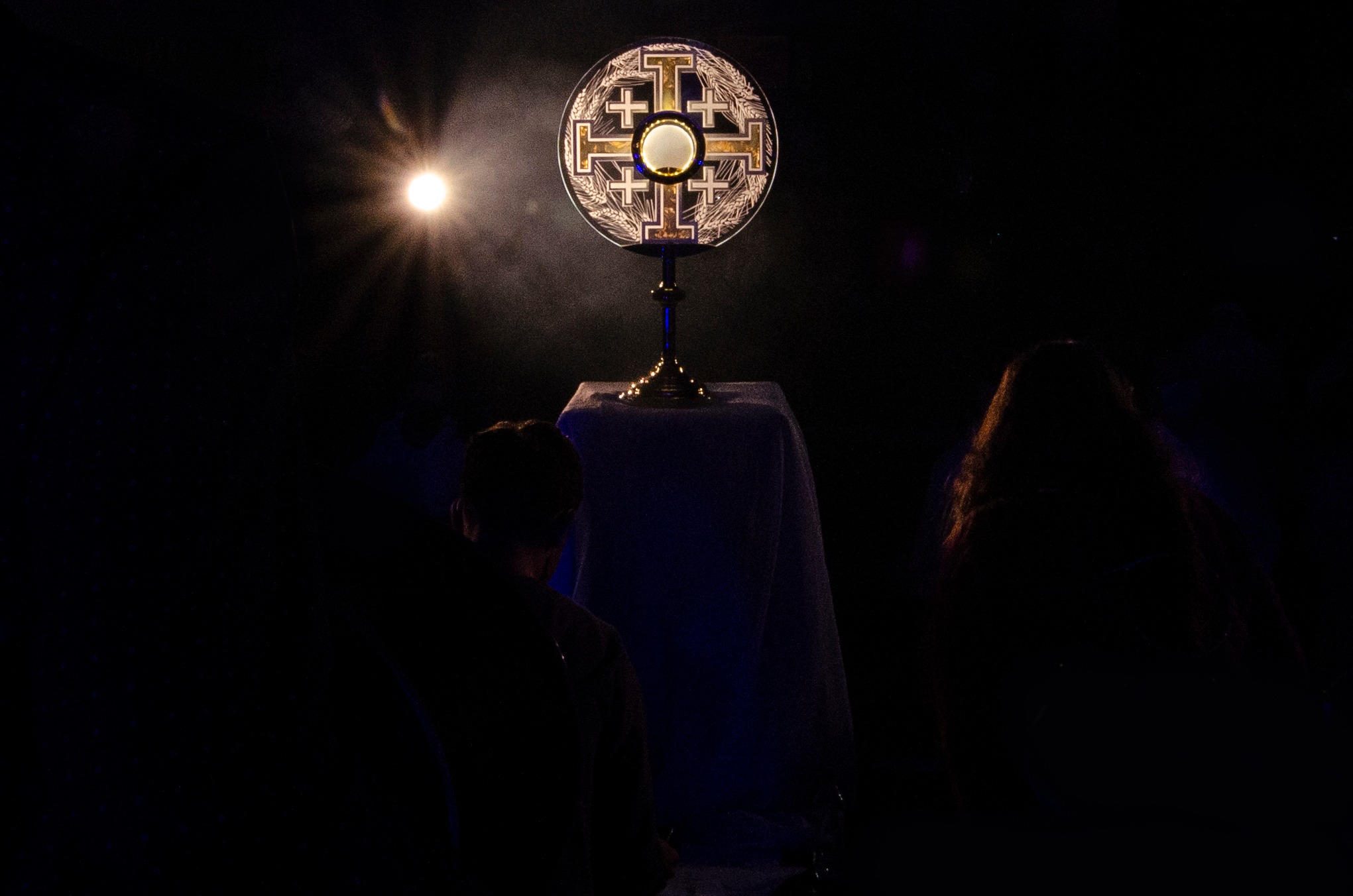 Monstrancja z Najświętszym Sakramentem na ołtarzu, oświetlona punktowym światłem, widziana zza pleców wiernych w zaciemnionym kościele.