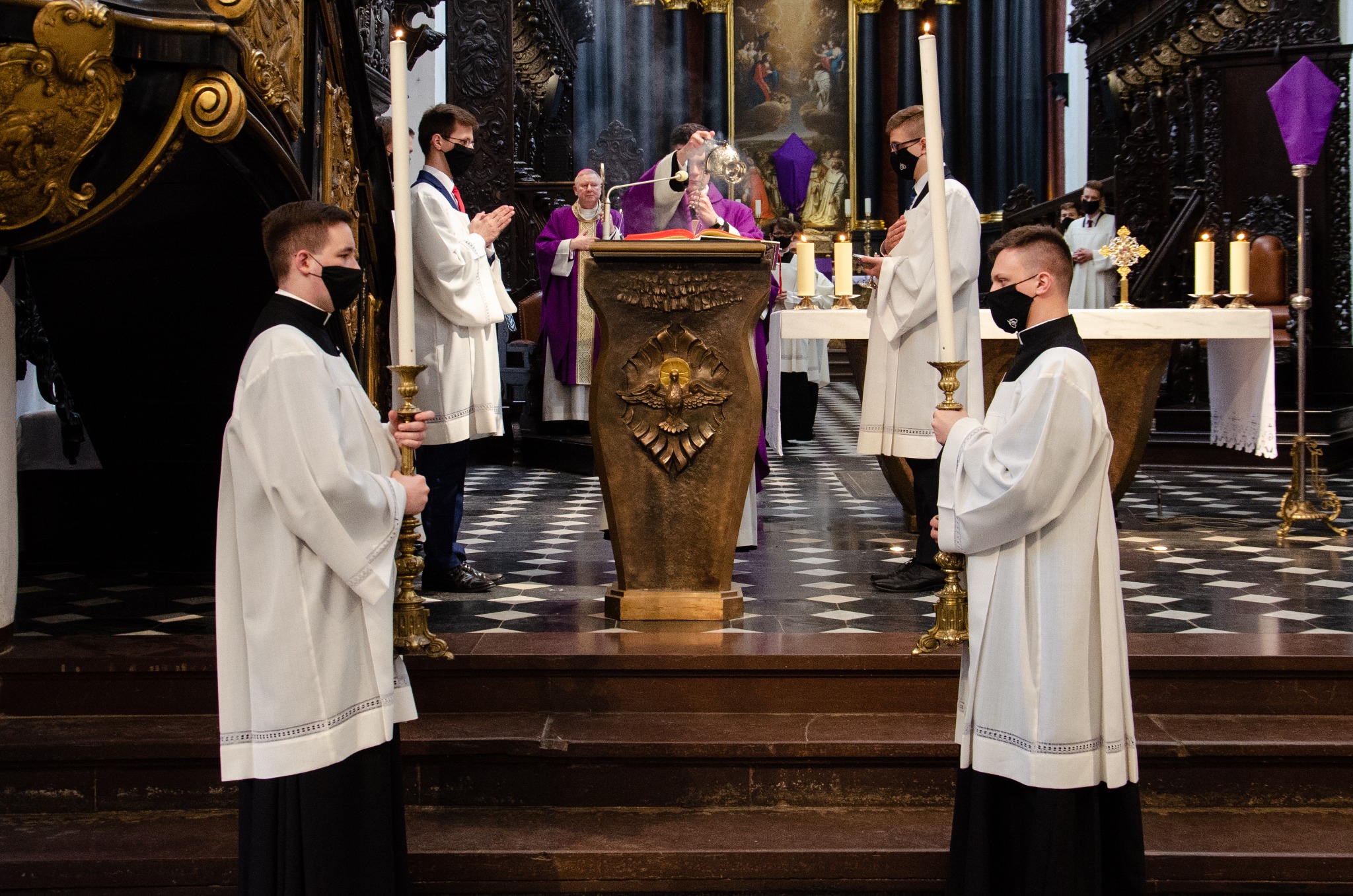 Wnętrze kościoła podczas mszy, ministranci w białych strojach i czarnych maskach trzymają świece, ksiądz w fioletowej szacie kadzi, w tle obraz religijny.
