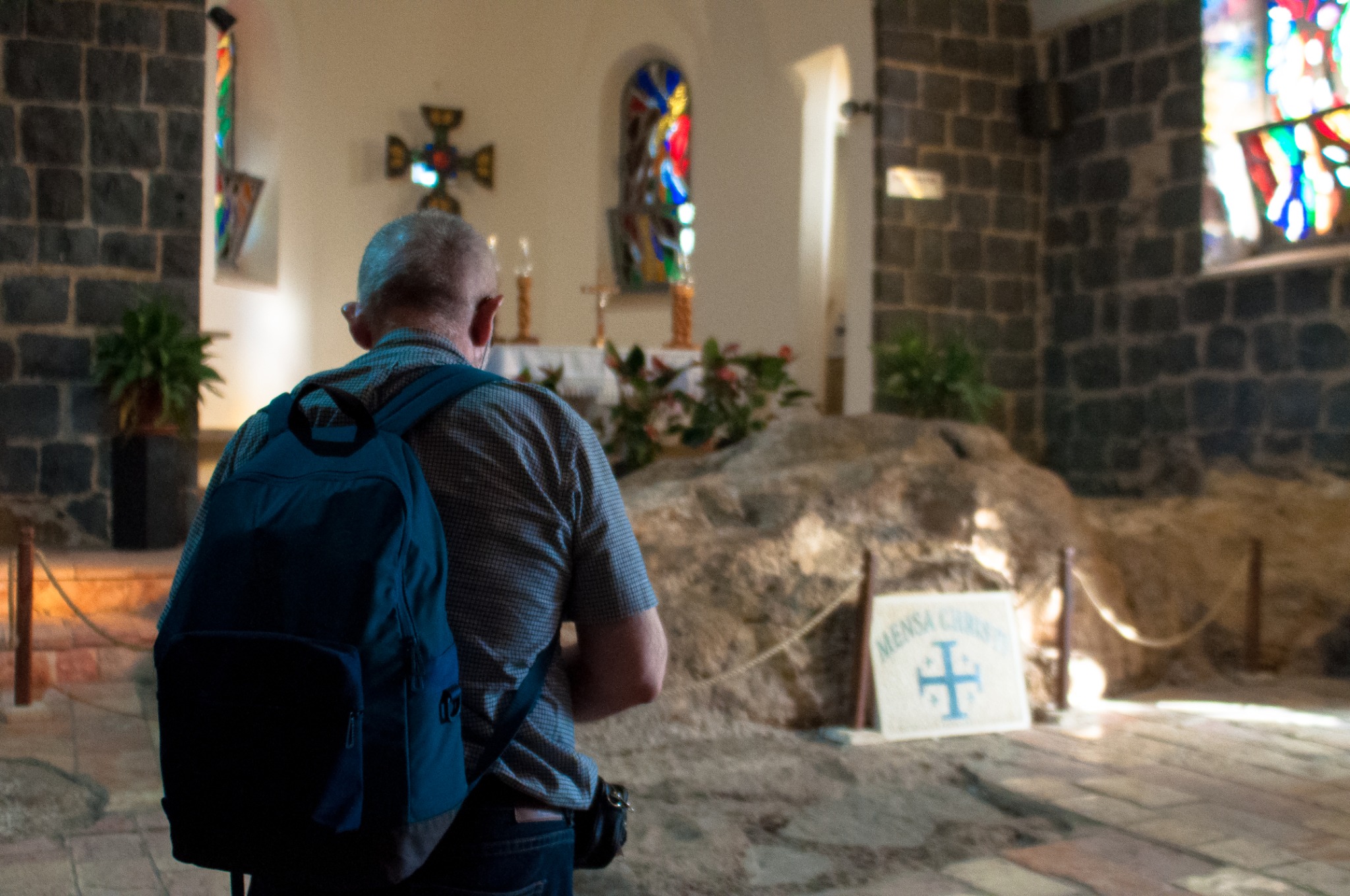 Mężczyzna z plecakiem i maseczką stoi w kościele Mensa Christi w Izraelu, patrząc na ołtarz z krzyżem i witraże.