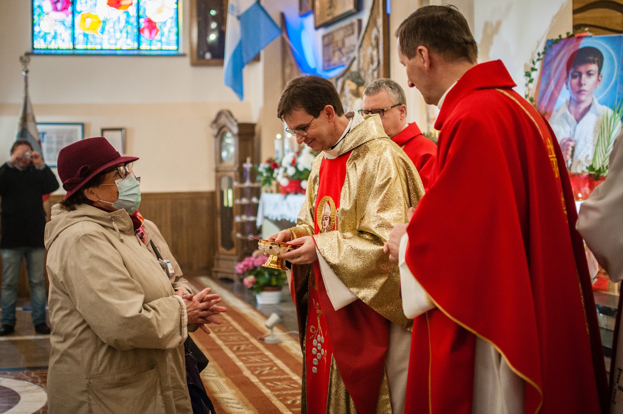 Wnętrze kościoła podczas ceremonii: ksiądz w złotej szacie podaje naczynie kobiecie w bordowym kapeluszu i masce, w tle drugi ksiądz w czerwonej szacie i osoba robiąca zdjęcie.