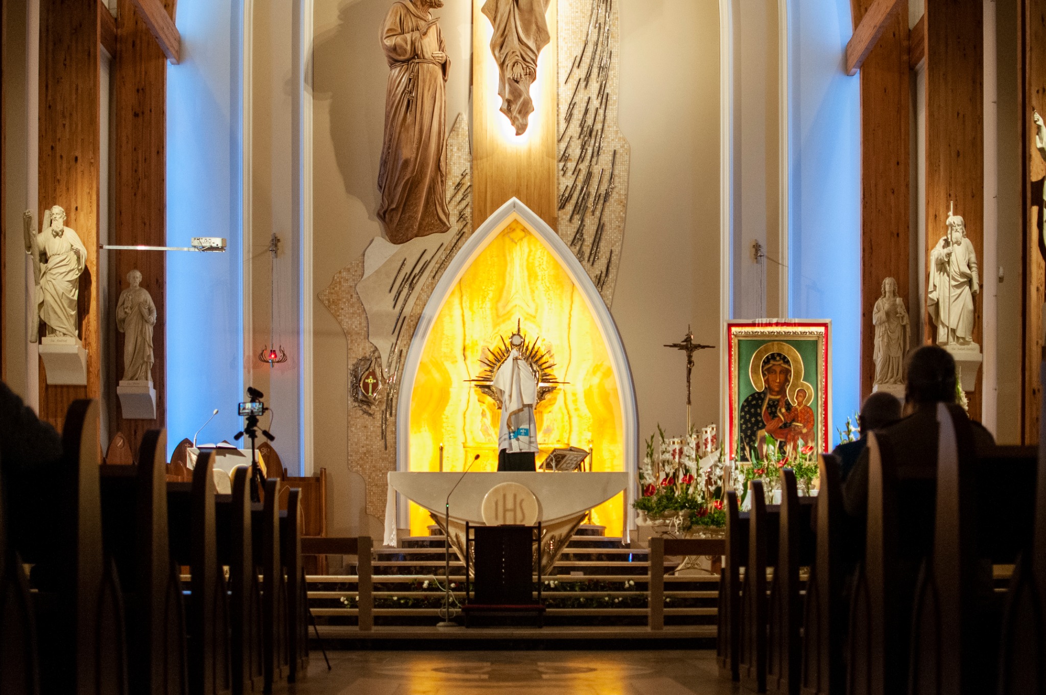 Wnętrze kościoła z ołtarzem podświetlonym na żółto, rzeźby świętych po bokach, ikona i krzyż widoczne z perspektywy nawy z rzędami ławek.