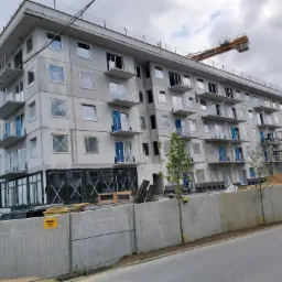 Wielopiętrowy budynek w trakcie budowy, z częściowo zamontowanymi oknami i balkonami, widoczne rusztowania i żuraw budowlany na tle pochmurnego nieba.