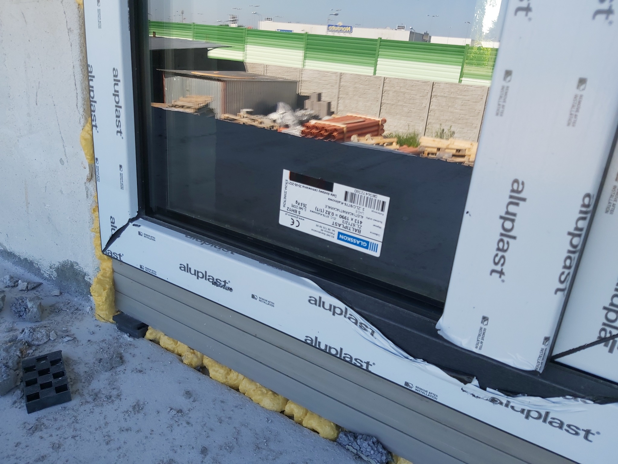 Fragment nowo zamontowanego okna z ramą oklejoną folią ochronną Aluplast, uszczelnionego pianką montażową, widoczne podkładki poziomujące i elementy placu budowy w tle.