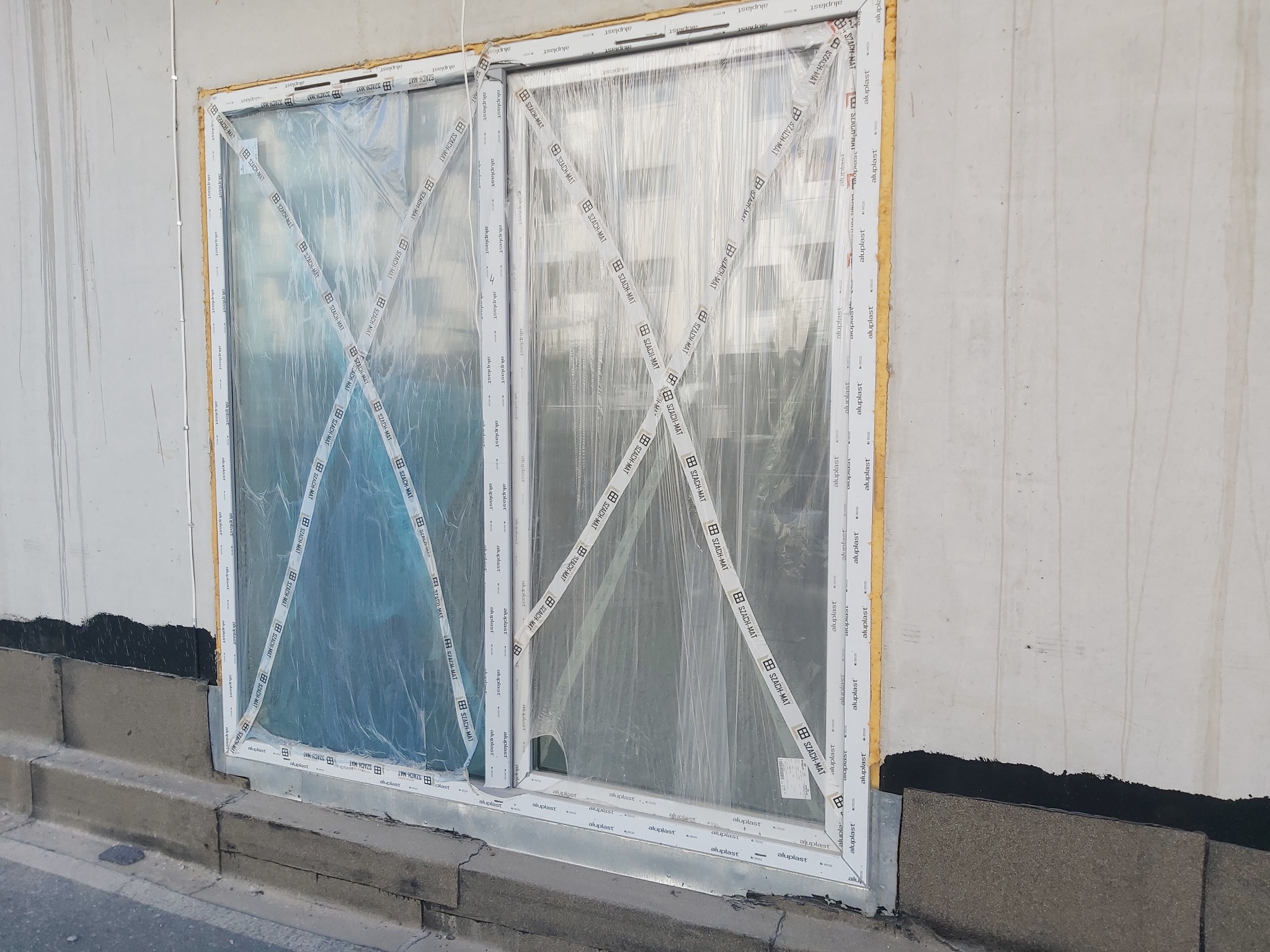 Nowo zamontowane, podwójne okno z białego profilu, zabezpieczone folią i taśmą ochronną z logo producenta na elewacji budynku w trakcie wykończenia.