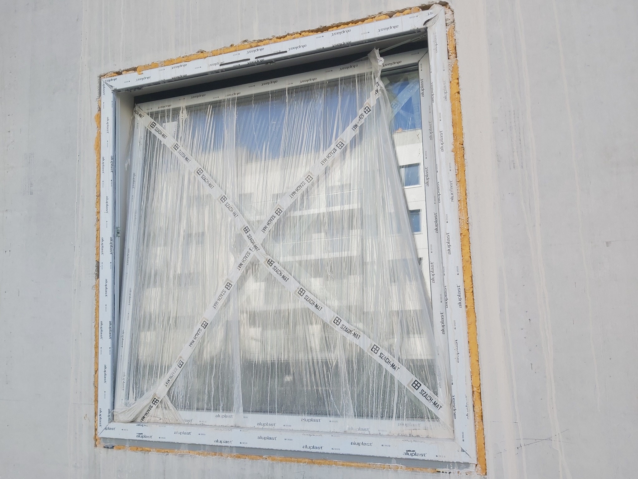 Nowo zamontowane okno z folią ochronną i taśmą zabezpieczającą SZACH-MAT, widoczne piana montażowa i surowy beton ściany.