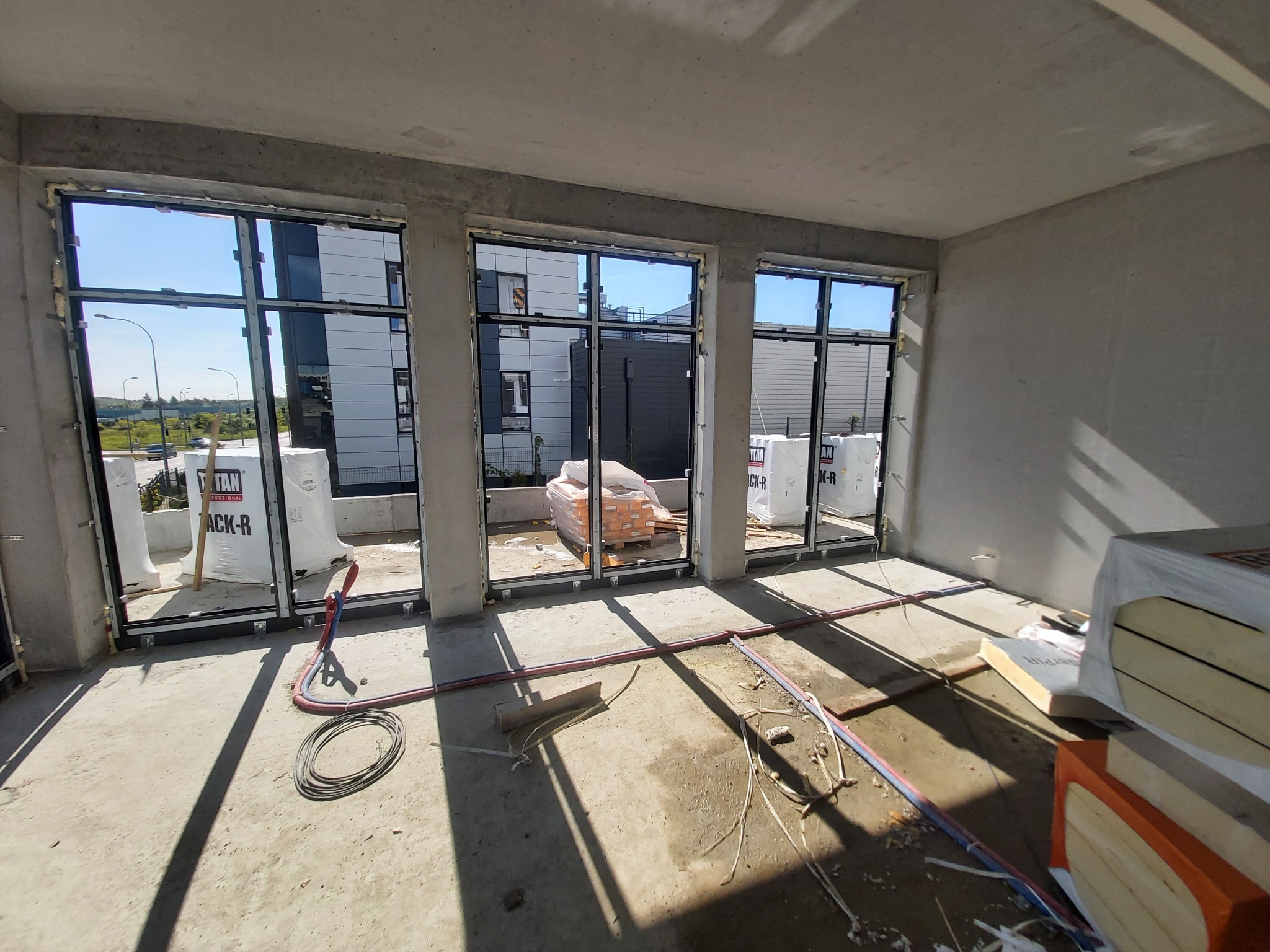 Surowy stan deweloperski: zamontowane okna aluminiowe z widokiem na budynki mieszkalne, materiały budowlane na podłodze, widoczne przewody instalacyjne.