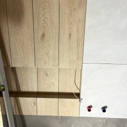 Baugrema Grzegorz Keczmerski - Montaż okładziny ściennej: połączenie paneli drewnopodobnych i białych płytek ceramicznych w łazience, widoczne przyłącza hydrauliczne i poziomica.