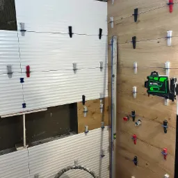 Układanie białych, falistych płytek na ścianie obok drewnopodobnych płytek, z użyciem poziomicy laserowej Hilda i systemu poziomowania płytek.