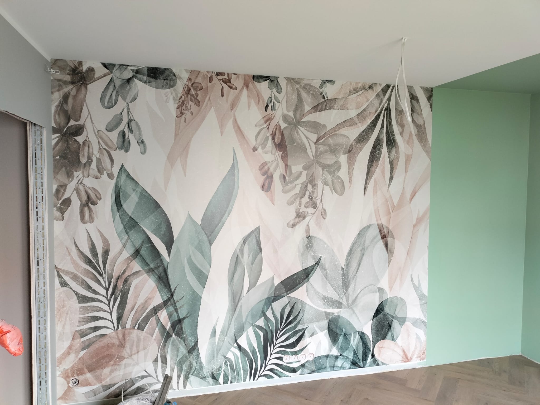 Ściana z fototapetą w motywy roślinne, sąsiadująca z pomalowaną na zielono ścianą, na podłodze parkiet ułożony w jodełkę.