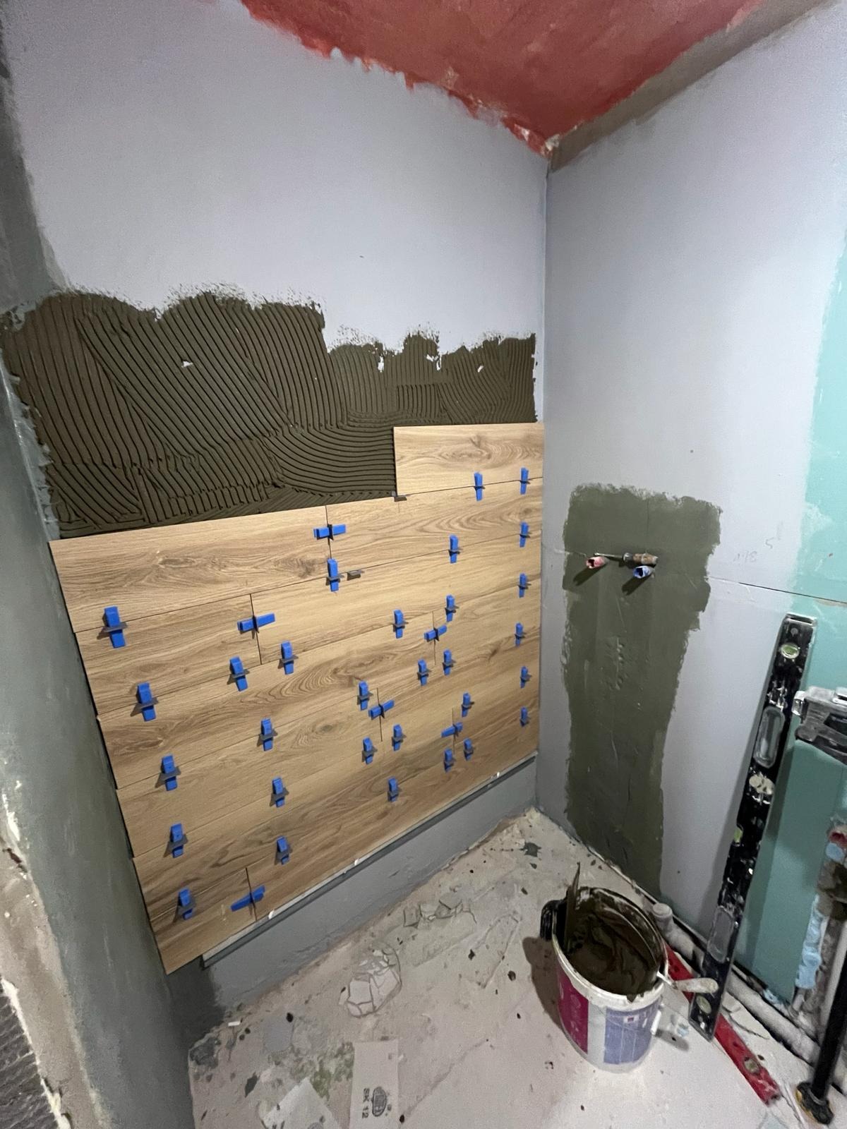 Ściana łazienki w trakcie układania płytek imitujących drewno, z widocznymi niebieskimi krzyżykami dystansowymi, klejem na ścianie i poziomnicą w rogu pomieszczenia.