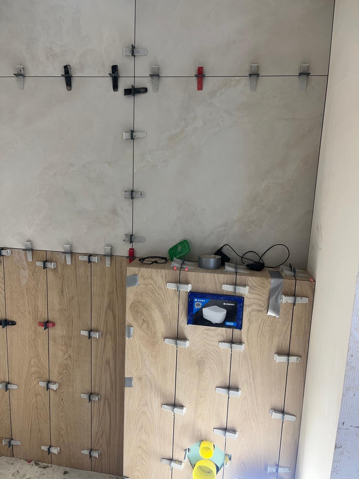 Układanie płytek imitujących drewno na ścianie, z systemem poziomowania płytek, widoczne narzędzia i opakowanie produktu Geberit.