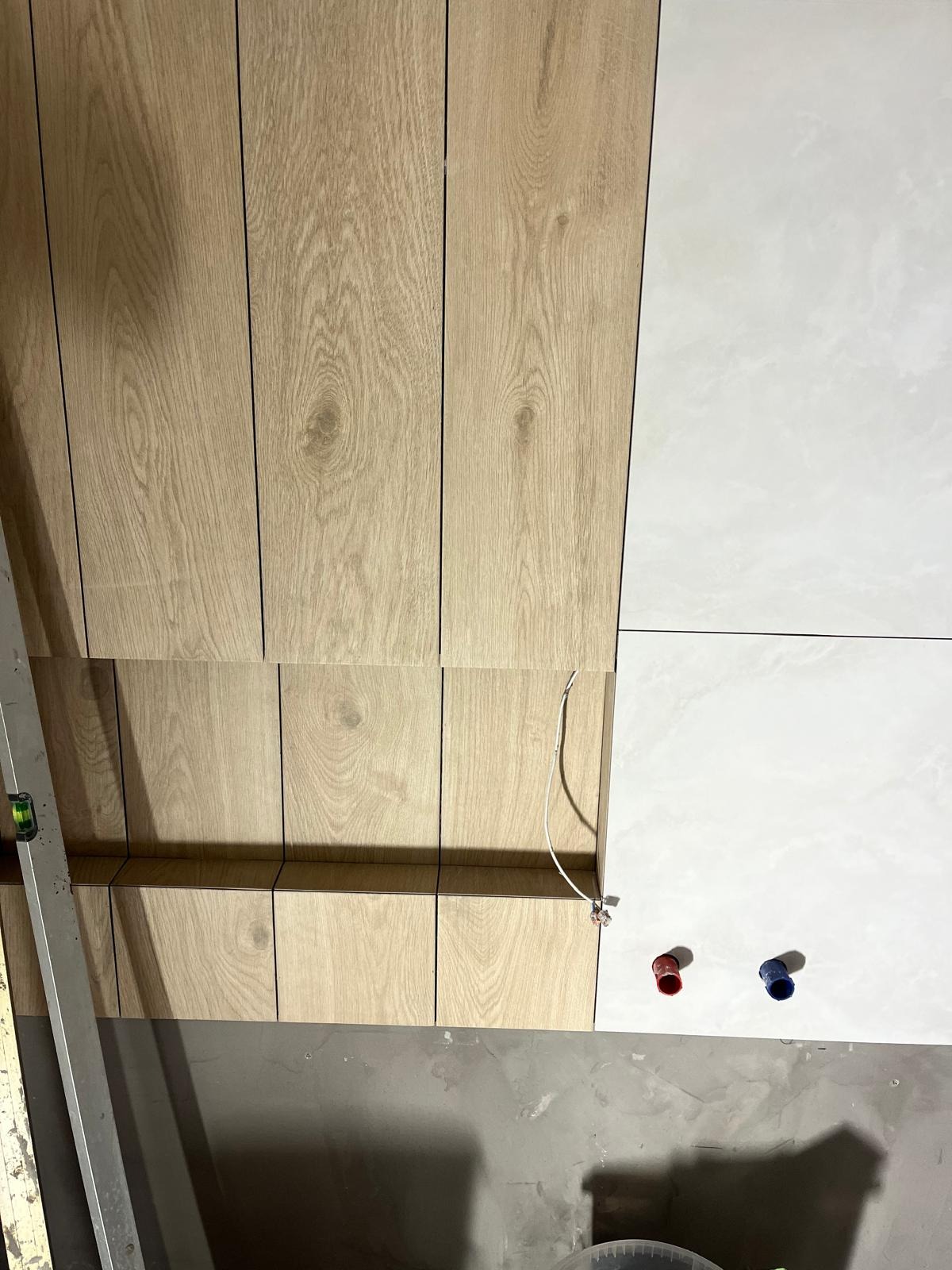 Montaż okładziny ściennej: połączenie paneli drewnopodobnych i białych płytek ceramicznych w łazience, widoczne przyłącza hydrauliczne i poziomica.