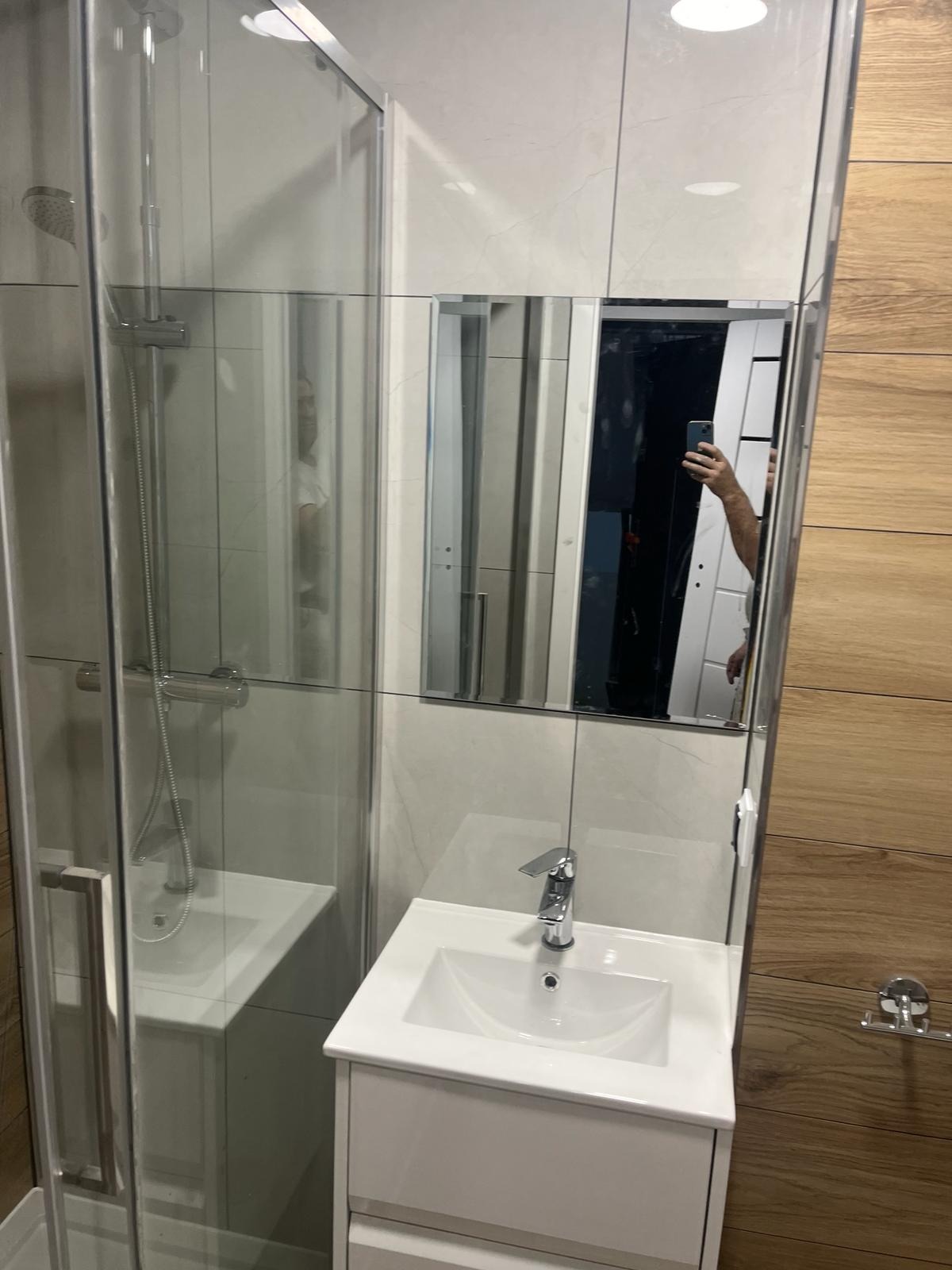 Nowoczesna łazienka po remoncie: kabina prysznicowa ze szklanymi drzwiami, biała umywalka z szafką, lustro z odbiciem fotografa i fragment ściany wyłożonej płytkami imitującymi drewno.