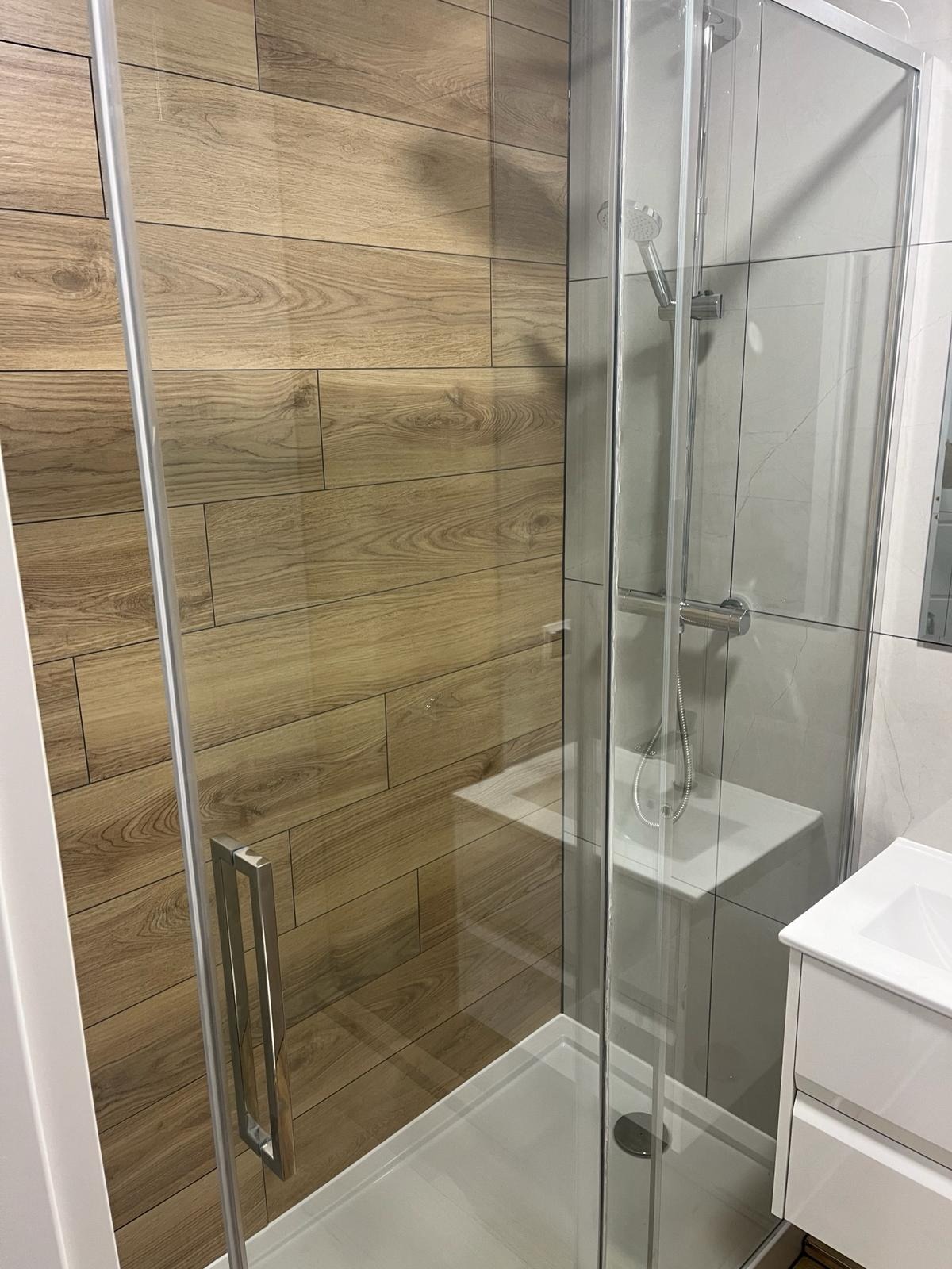 Kabina prysznicowa z drewnopodobnymi płytkami na ścianie i szarymi płytkami imitującymi marmur, biały brodzik i umywalka z szafką.