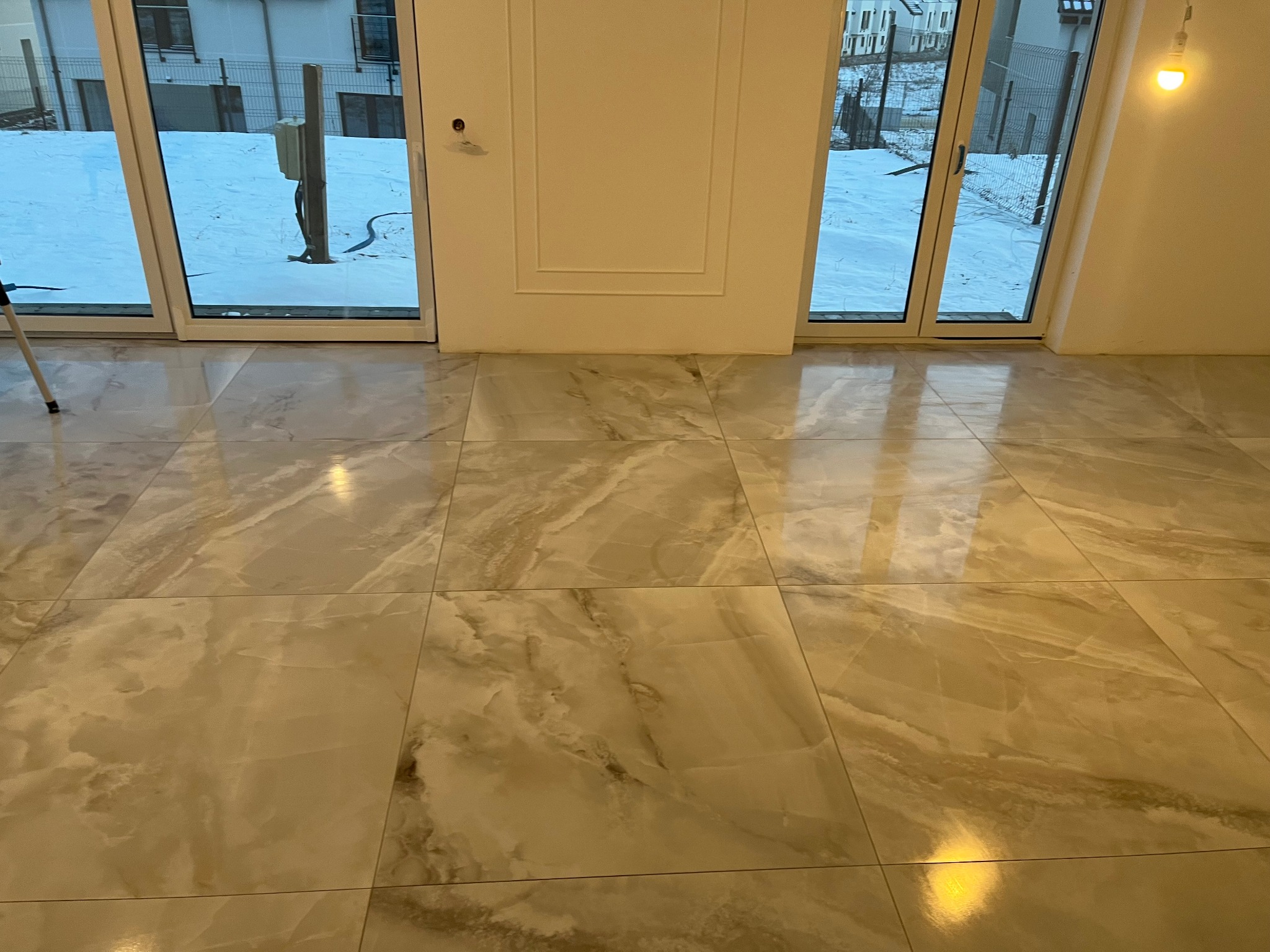Beżowa, marmurkowa podłoga z płytek ceramicznych, świeżo położona w jasnym pomieszczeniu z dużymi oknami balkonowymi, przez które widać śnieg.