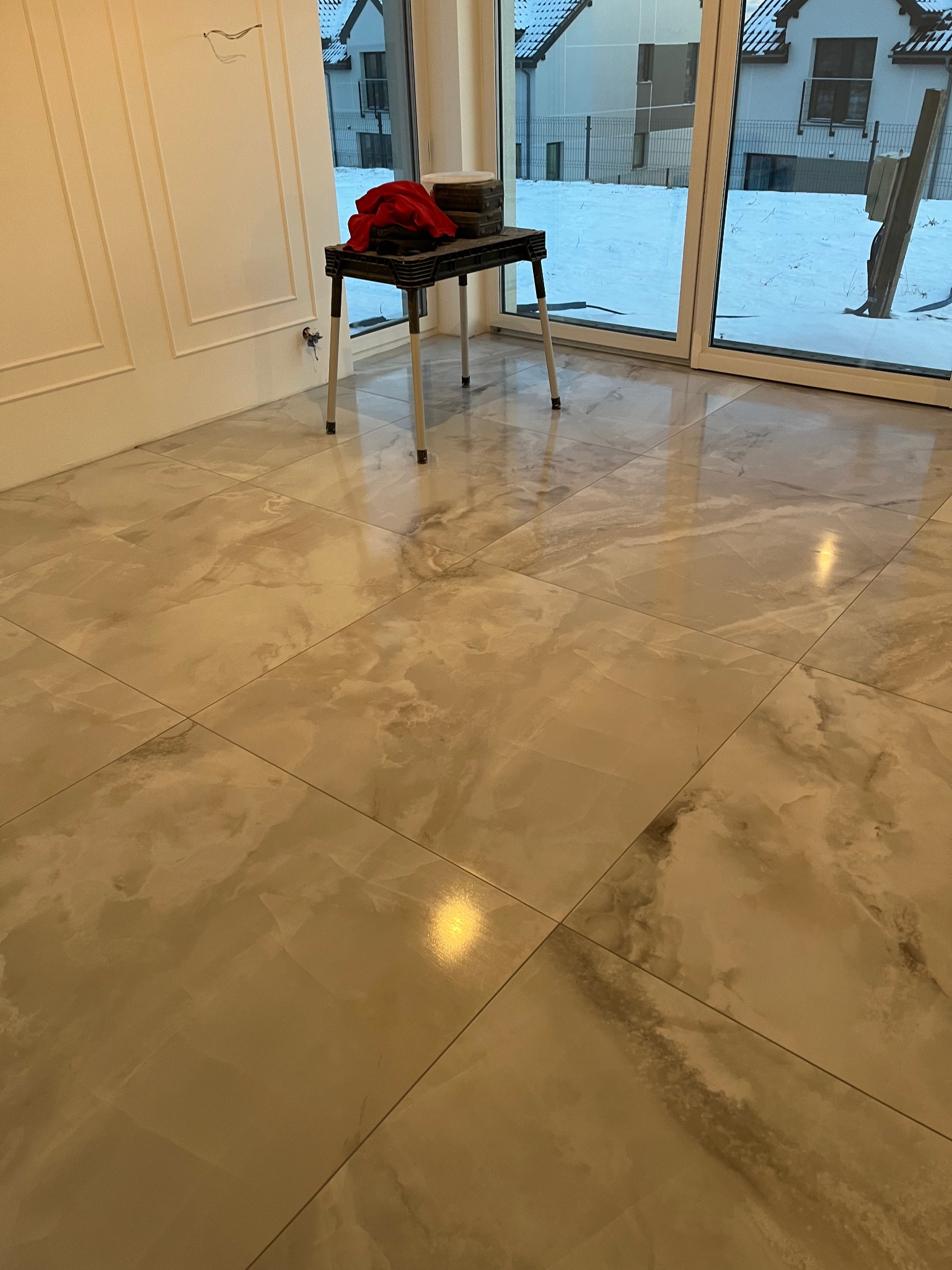 Beżowe płytki podłogowe imitujące marmur, widoczne okno z widokiem na ośnieżony ogród i przygotowane narzędzia na małym stoliku.