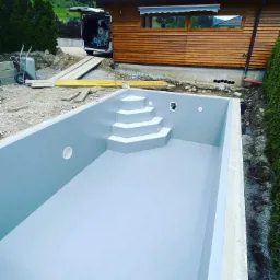 Wnętrze nowo wybudowanego, prostokątnego basenu z wbudowanymi schodami w Czudcu, widoczne przygotowania do dalszych prac wykończeniowych, w tle budynek z drewnianą elewacją i samochód dostawczy...