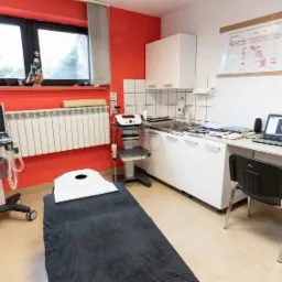 Gabinet fizjoterapeutyczny z leżanką pokrytą ciemnym materiałem, aparaturą do fizykoterapii na mobilnym stoliku, biurkiem z laptopem i model anatomiczny człowieka, ściana pomalowana częściowo...