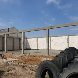 Surowy stan budowy domu z betonu kom&oacute;rkowego, widoczne ściany, betonowe słupy i wylany fundament, na pierwszym planie sterta opon od maszyn rolniczych.