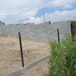 Nowo postawiony, szary, betonowy płot panelowy z fakturą kamienia, widok z zewnątrz, z widocznymi słupkami montażowymi i przygotowanym podłożem z ziemi.