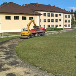 Żółta koparka Liebherr ładuje ziemię do czerwonej ciężarówki na tle budynku szkolnego i zielonego boiska. Widoczne są ślady po pracach ziemnych na ścieżce.