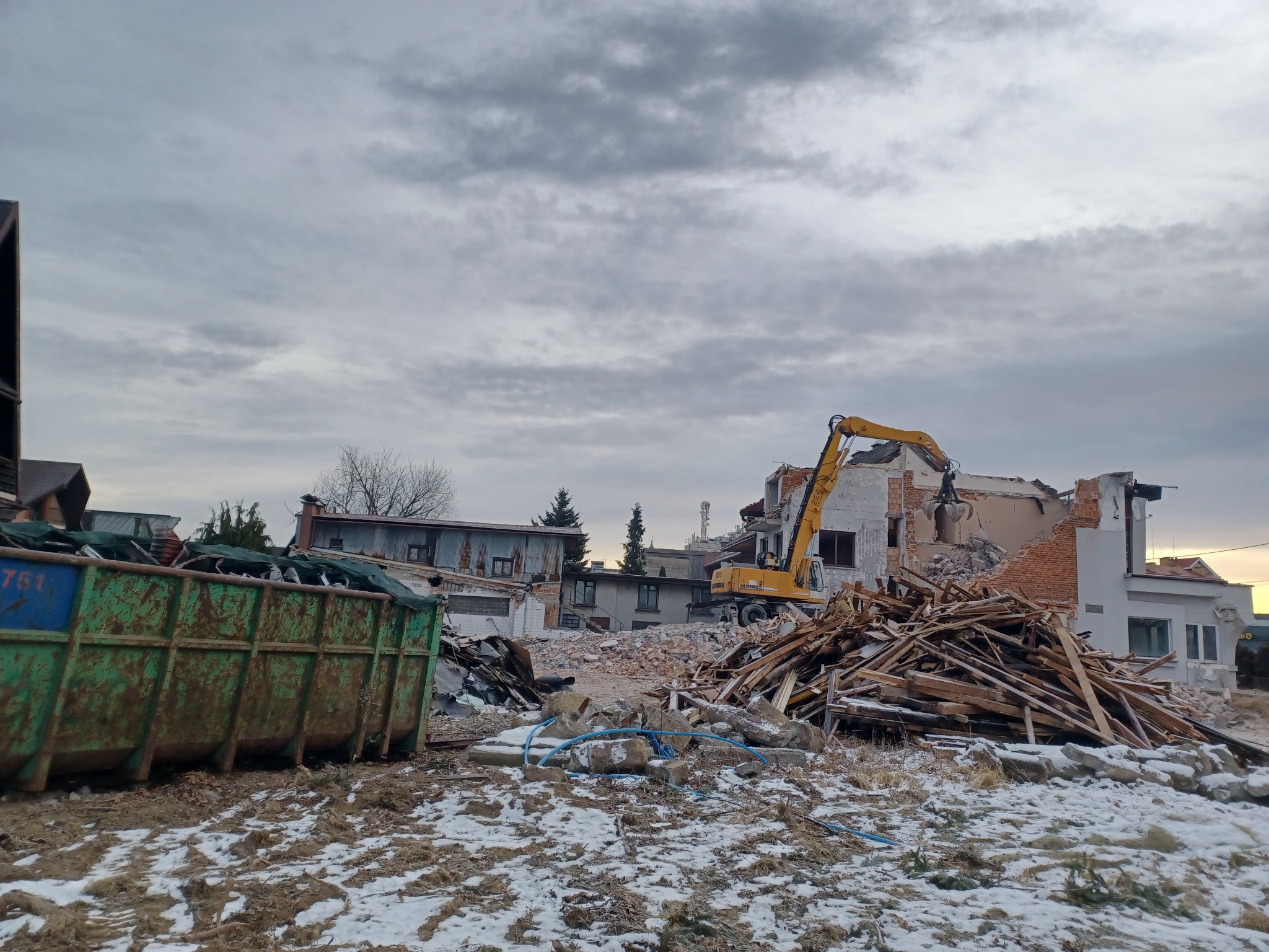 Żółta koparka Liebherr z chwytakiem na tle częściowo rozebranego budynku, gruzowisko, zielony kontener na śmieci, resztki śniegu na ziemi, pochmurne niebo.