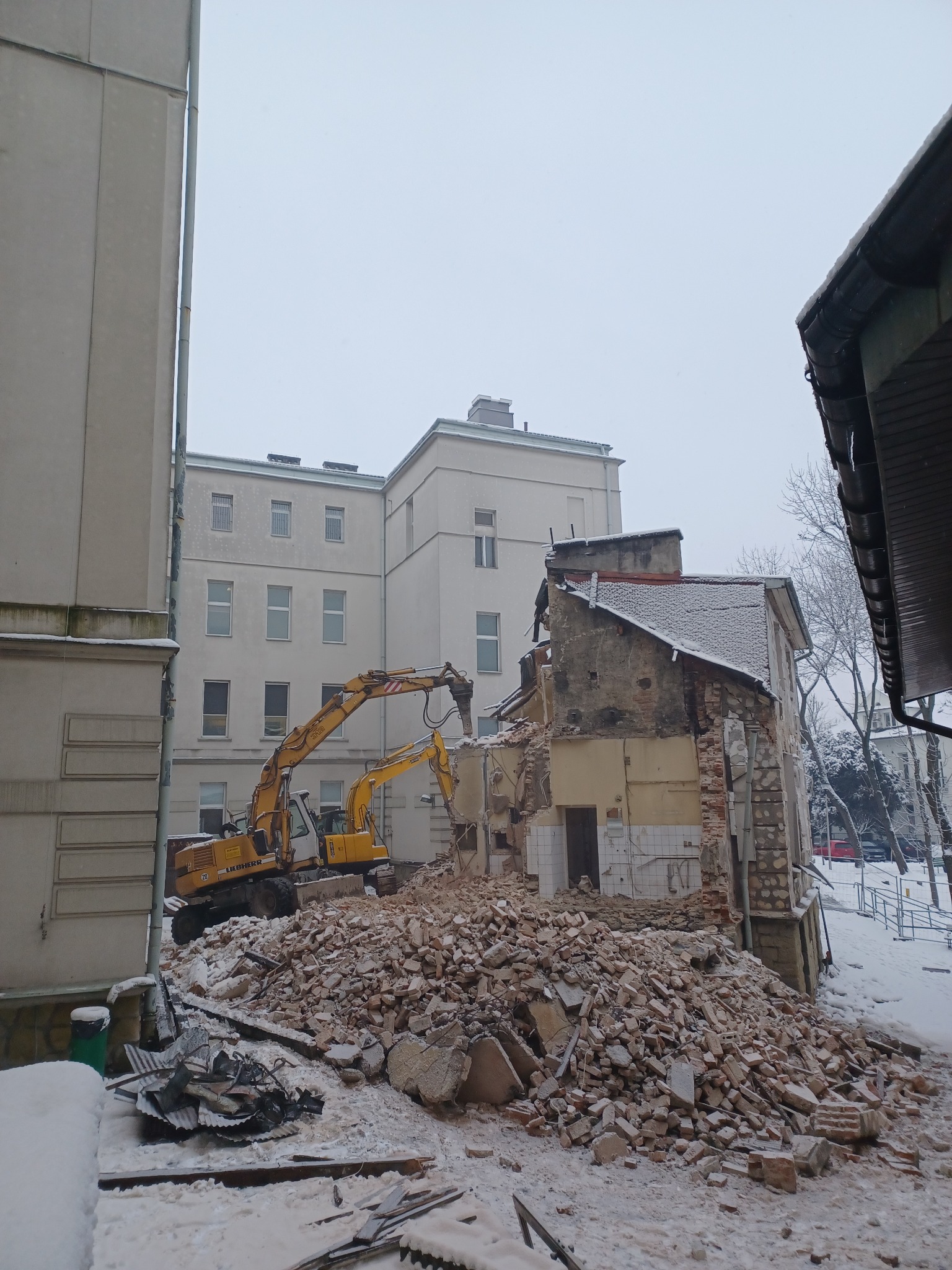 Żółty koparko-ładowarka Liebherr kruszy pozostałości budynku w zimowej scenerii, z gruzem i śniegiem w pierwszym planie, a w tle widać większy budynek.