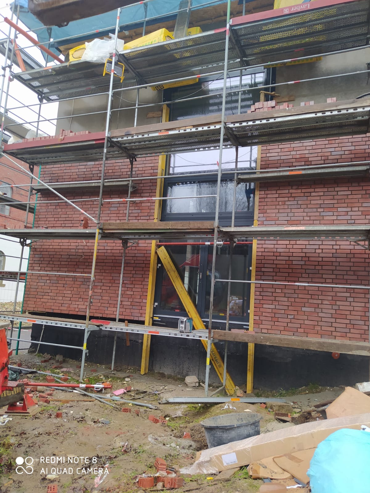 Wznoszenie ściany z cegły klinkierowej na budynku, widok z poziomu gruntu na rusztowanie z resztkami zaprawy, fragmenty cegieł na ziemi, okna z czarną ramą.