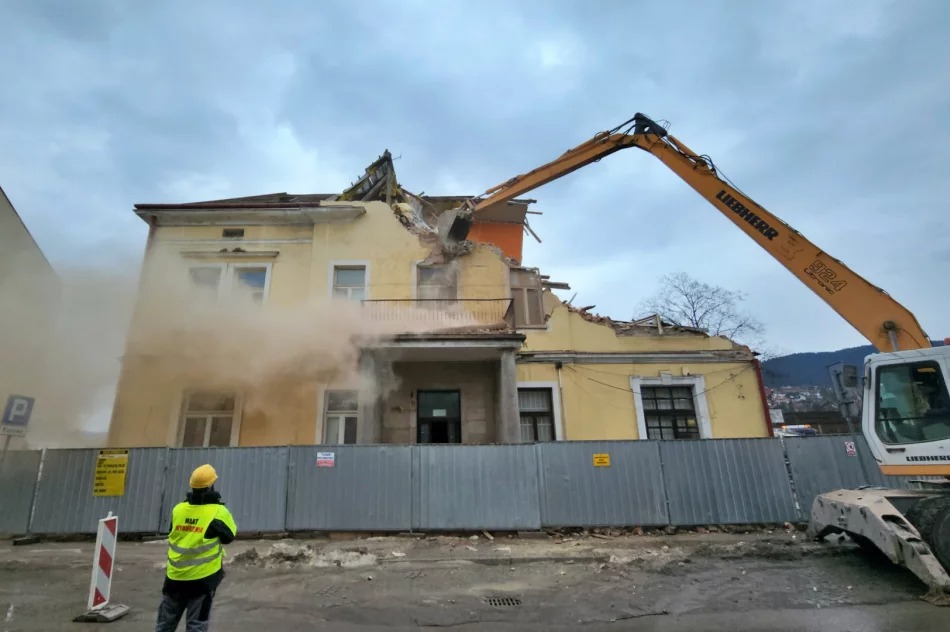 Żółty budynek w trakcie rozbiórki przez koparkę Liebherr 924, z widocznym pyłem i pracownikiem w żółtej kamizelce. Częściowo zniszczony dach i elewacja.