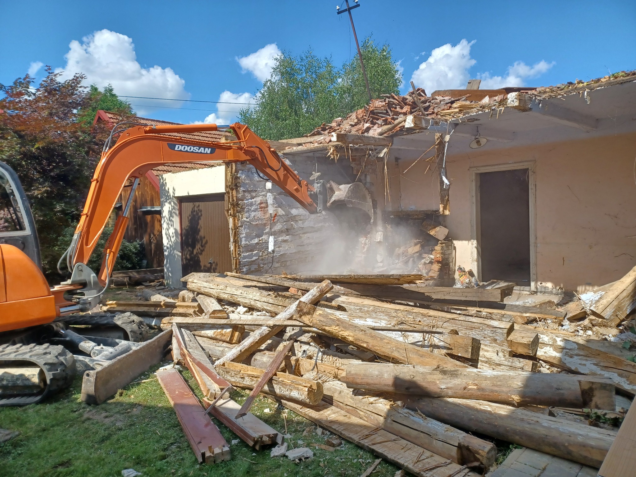 Pomarańczowa koparka Doosan w trakcie rozbiórki murowanego budynku w Nowym Sączu, z widocznymi drewnianymi belkami i gruzem. Kurz i pył unoszą się w powietrzu.